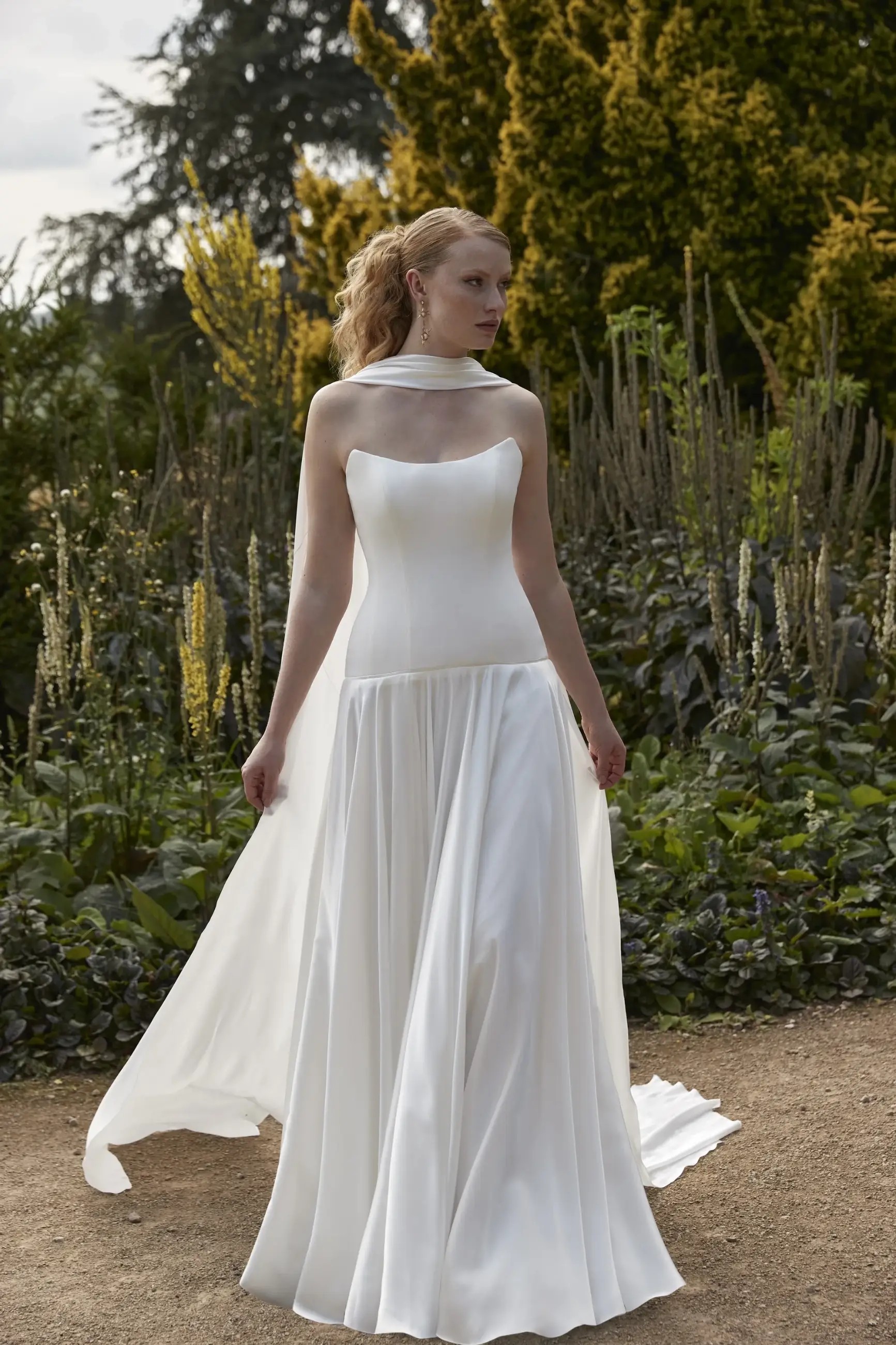 Model is wearing a strapless dropped waist wedding dress with a flowy skirt, structured bodice, and a chiffon neck scarf draped around her neck