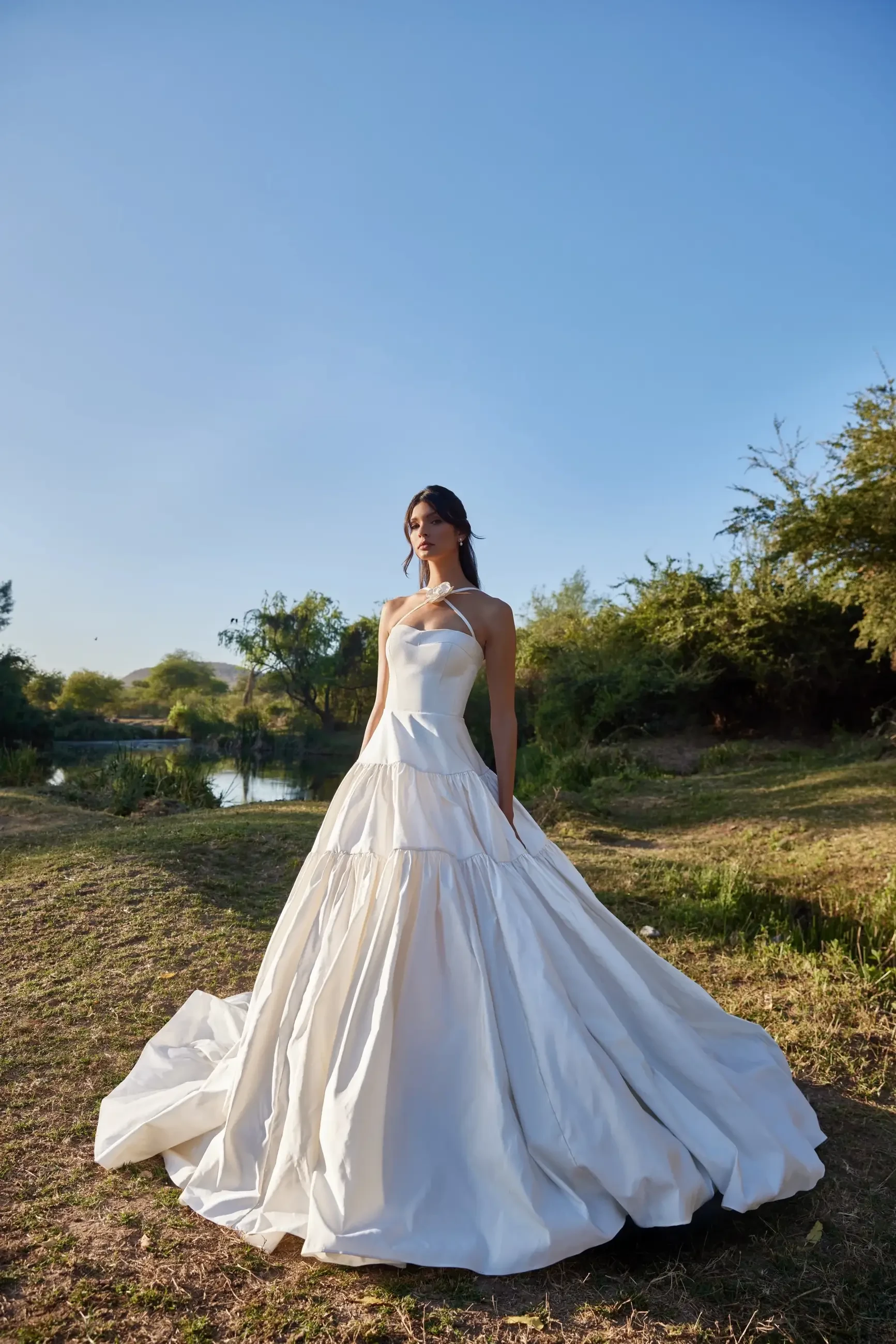 A woman wearing a white wedding gown stands in a grassy area with trees and a body of water in the background.