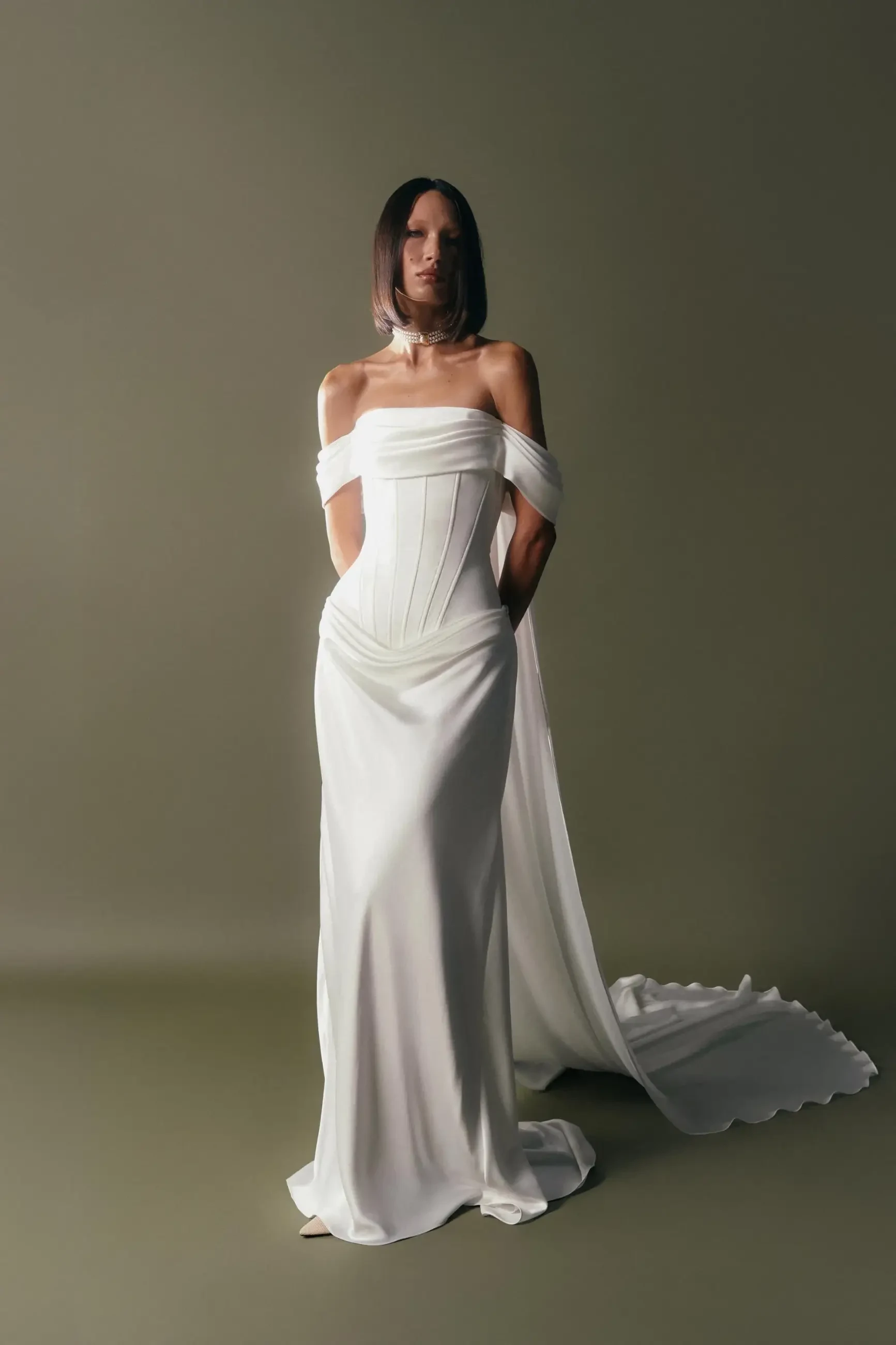 Model is wearing an off the shoulder satin bridal gown with a drop waist corset and cowl accent on the straight neckline 