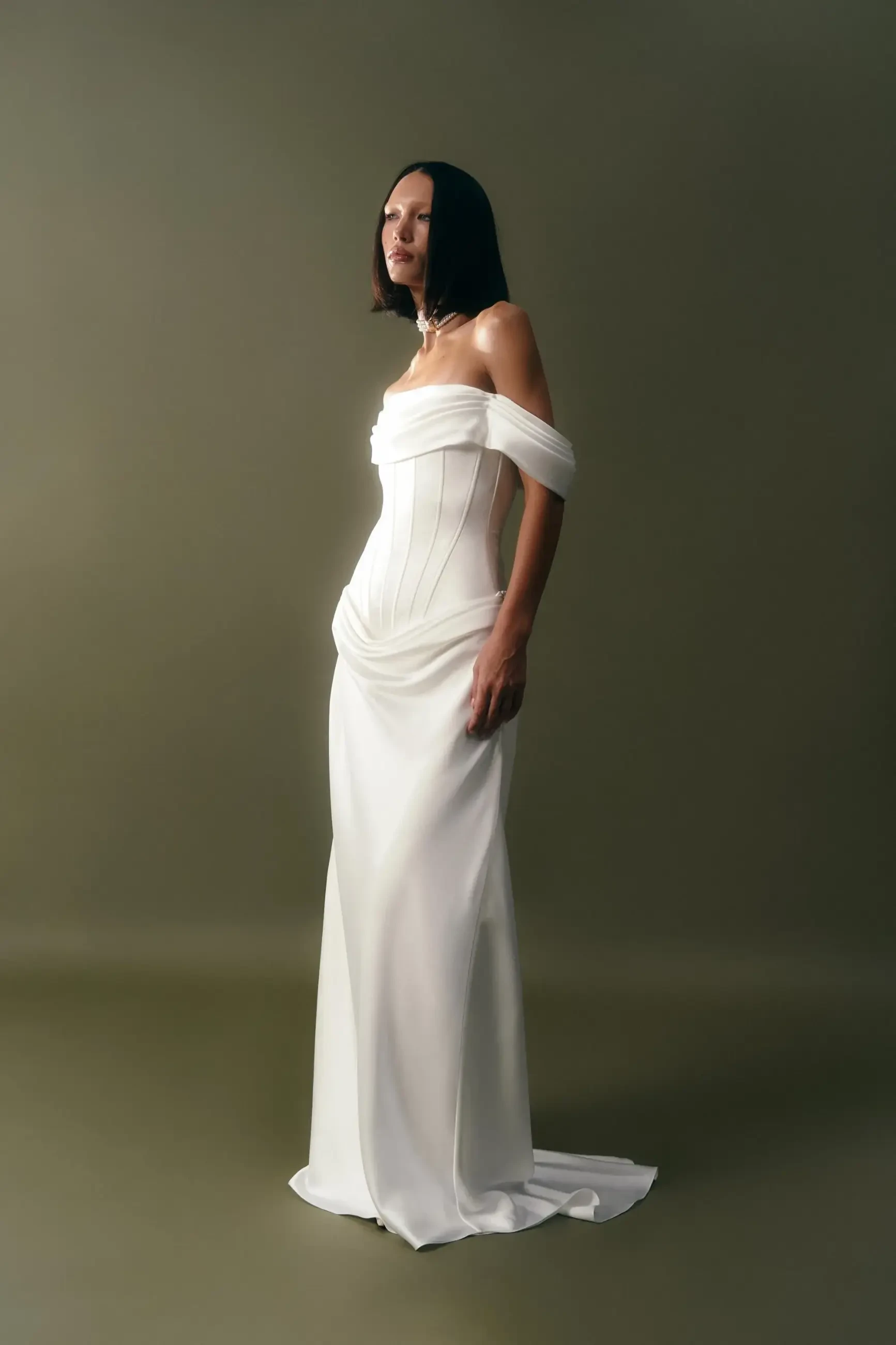 A model wearing a sleek, off-the-shoulder white gown, posing against a simple gray background.