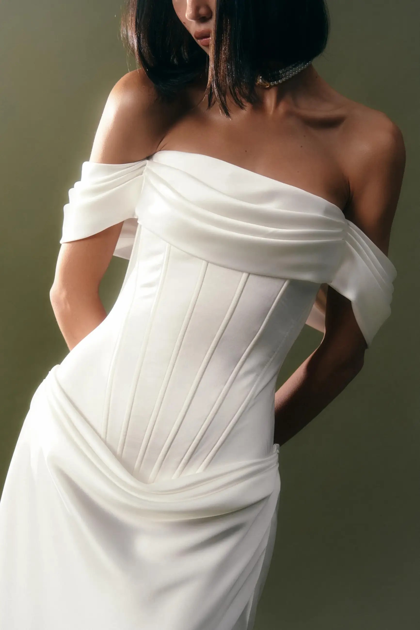 Model is wearing an off the shoulder wedding dress with a column silhouette and dropped waist with ruching on the bodice and hips