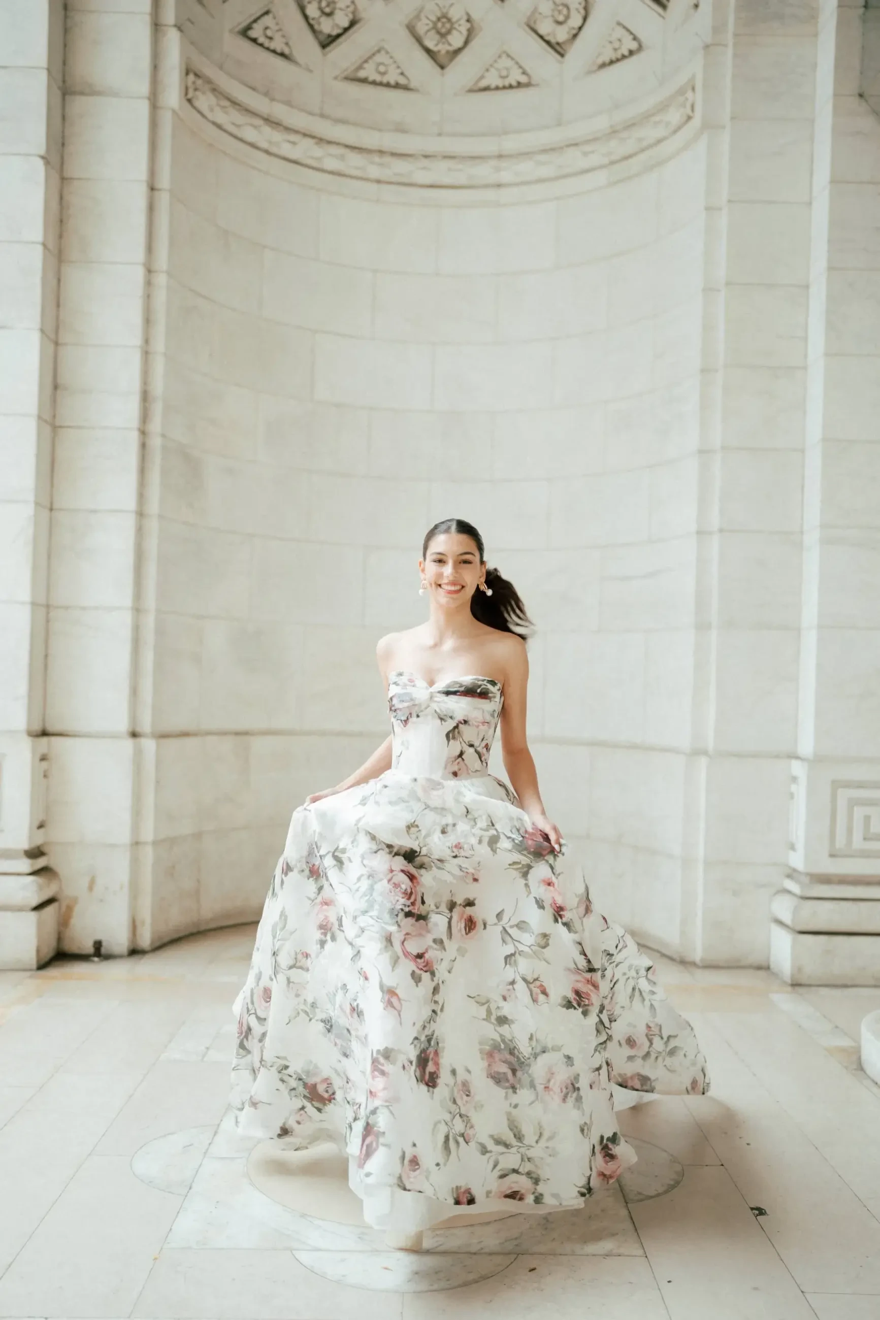 A woman in a floral-patterned gown stands gracefully in an elegant architectural space.