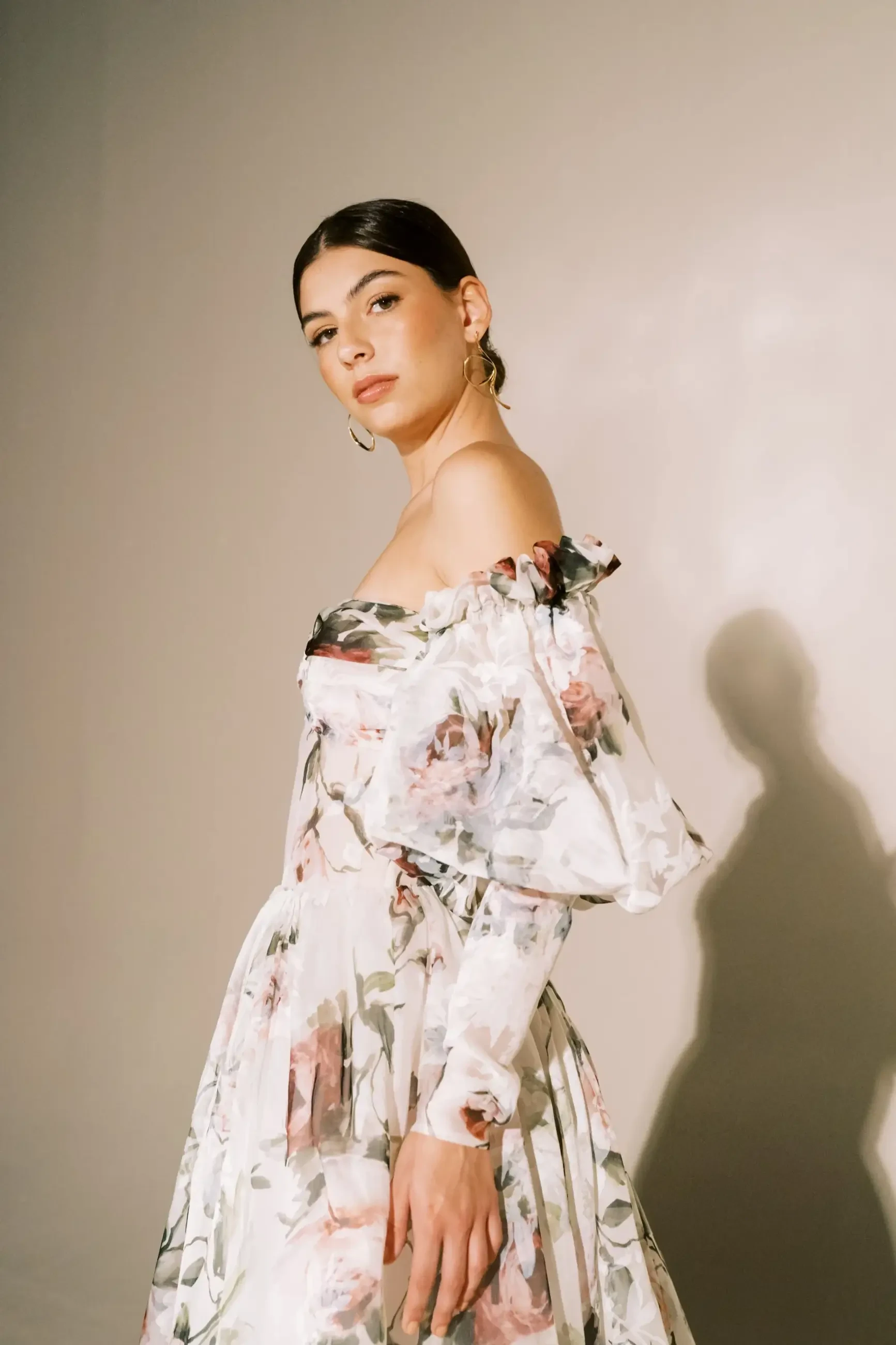 A woman in a floral off-shoulder dress stands against a neutral background, looking towards the camera with confidence.