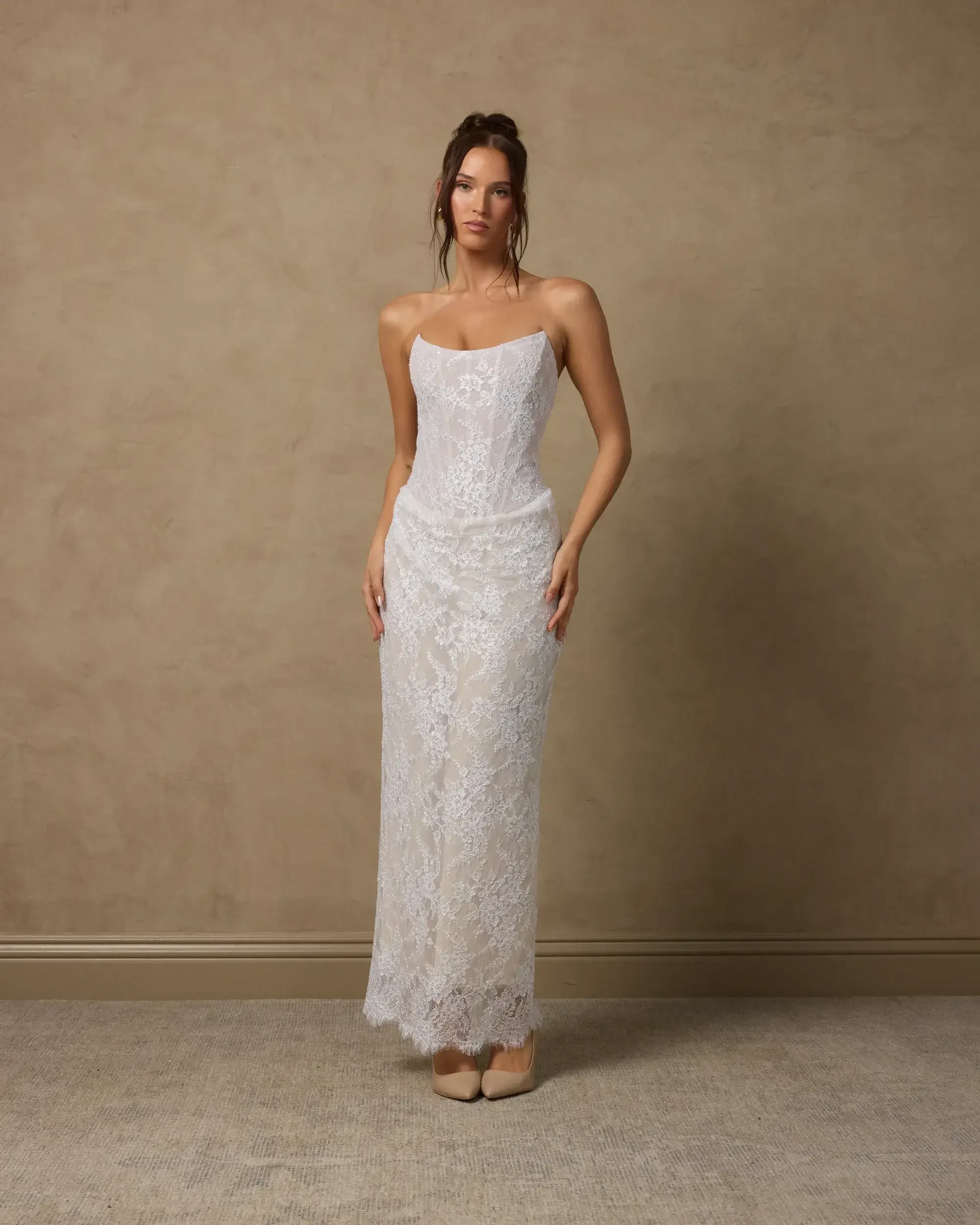 A woman stands in a fitted white lace dress against a neutral backdrop.