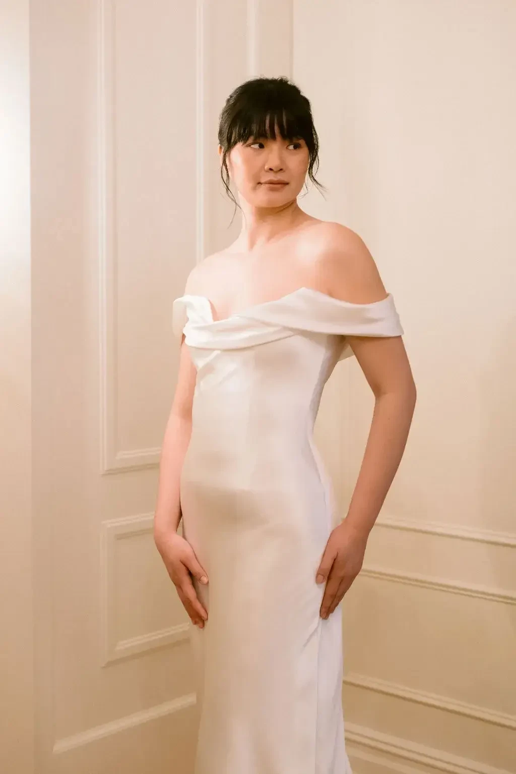 A woman in an elegant off-the-shoulder white dress stands in a softly lit room with light-colored walls.
