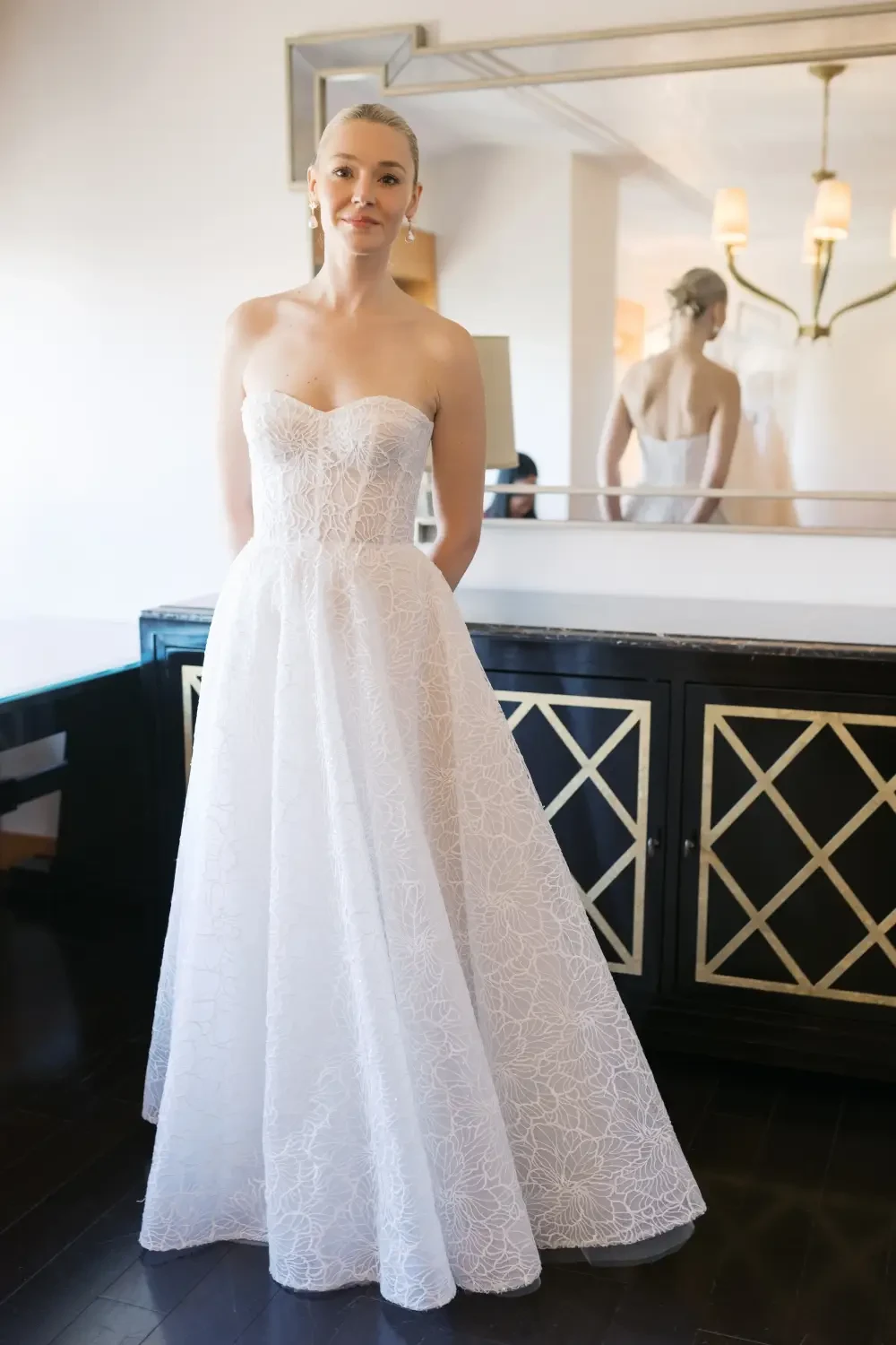 A woman in a strapless, lace wedding gown stands in a stylish room with a mirror behind her.