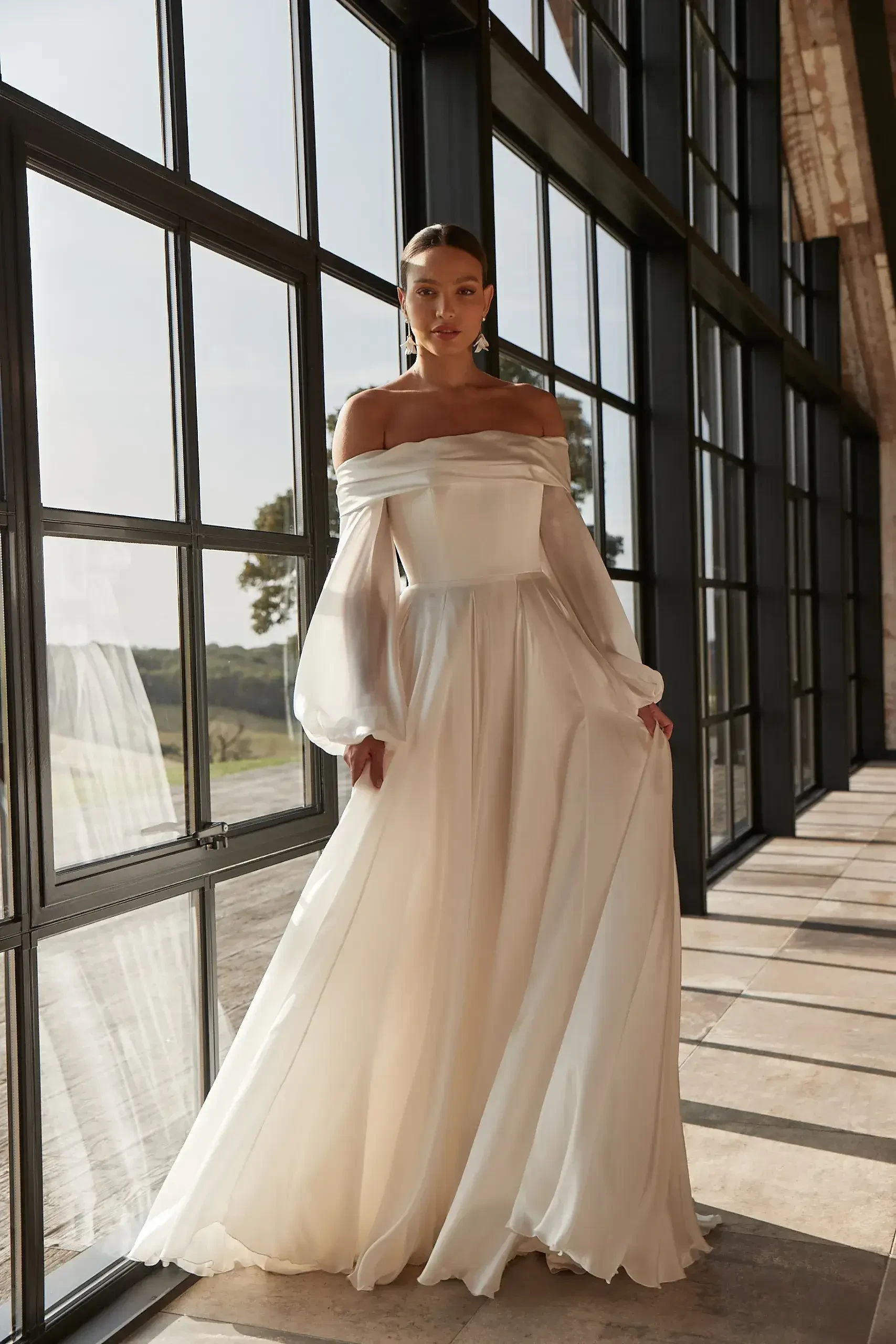 A woman in an elegant off-shoulder white gown stands by large windows, natural light highlighting her serene pose. The setting conveys a calm, graceful ambiance.