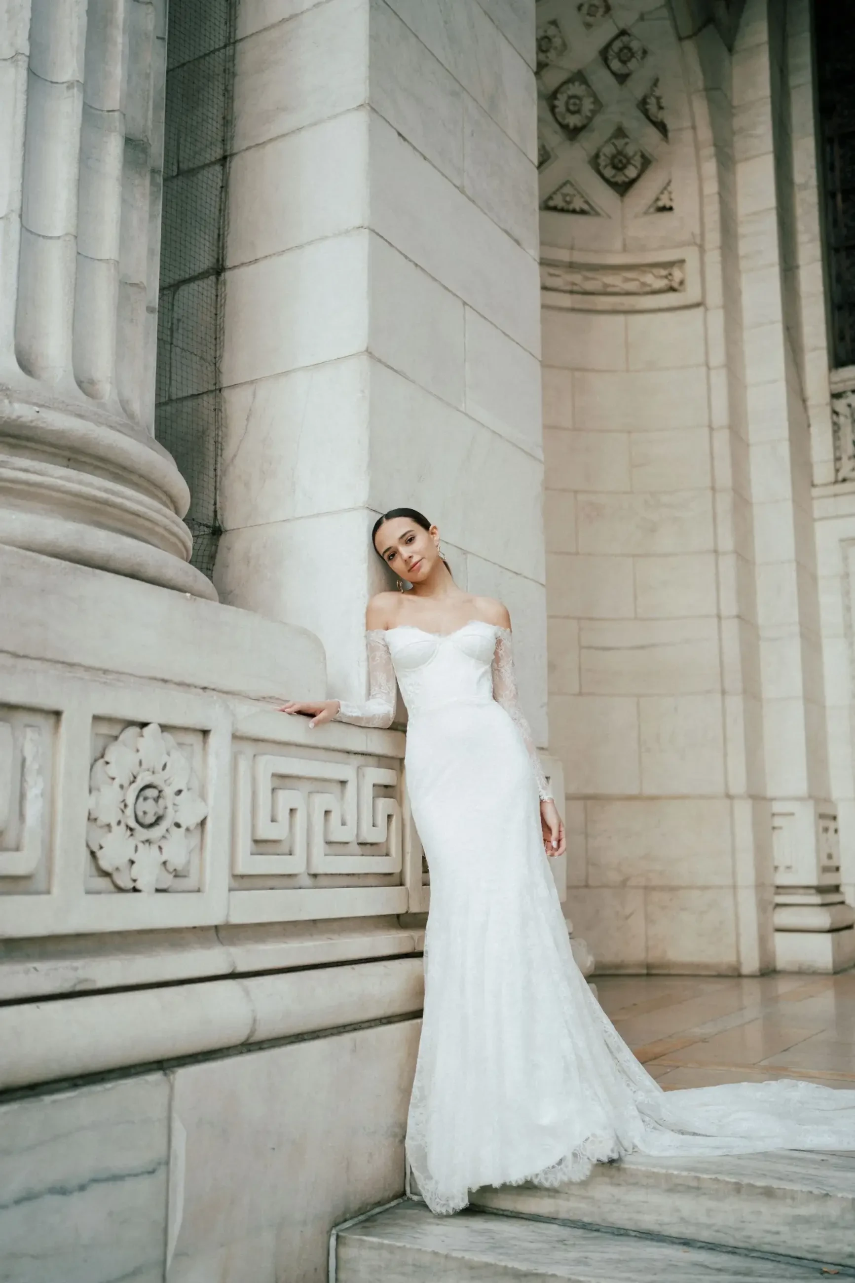 A bride in a white off-the-shoulder wedding gown poses against a marble column, with intricate architectural details in the background.