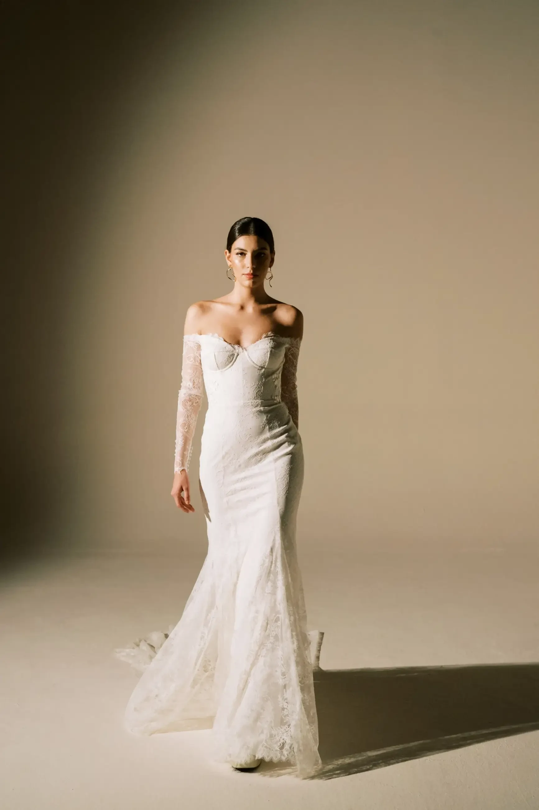 A woman in an elegant off-shoulder, long-sleeved lace wedding gown walks confidently on a beige background, exuding a serene and poised demeanor.