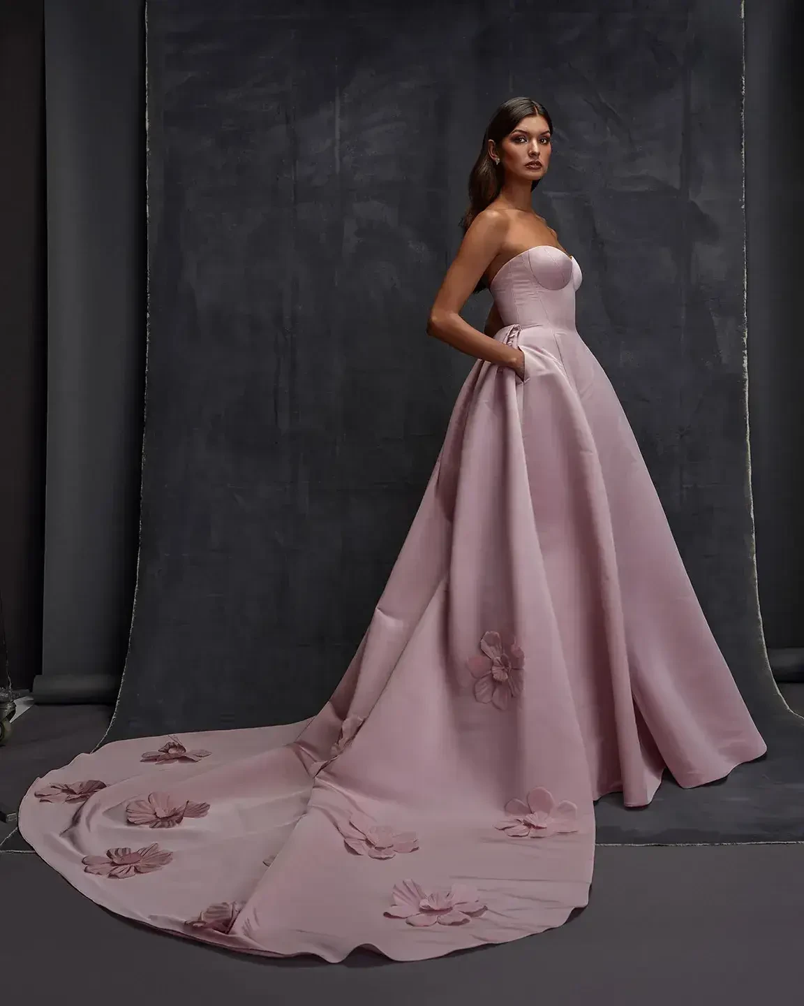 Model is wearing a strapless light pink wedding dress with a sculpted bodice and long train with large floral accents 