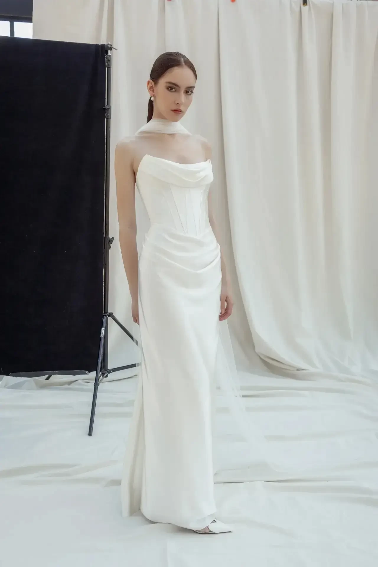 A model wearing a white strapless gown stands against a neutral backdrop with soft lighting. The gown features draped fabric that elegantly flows to the floor.