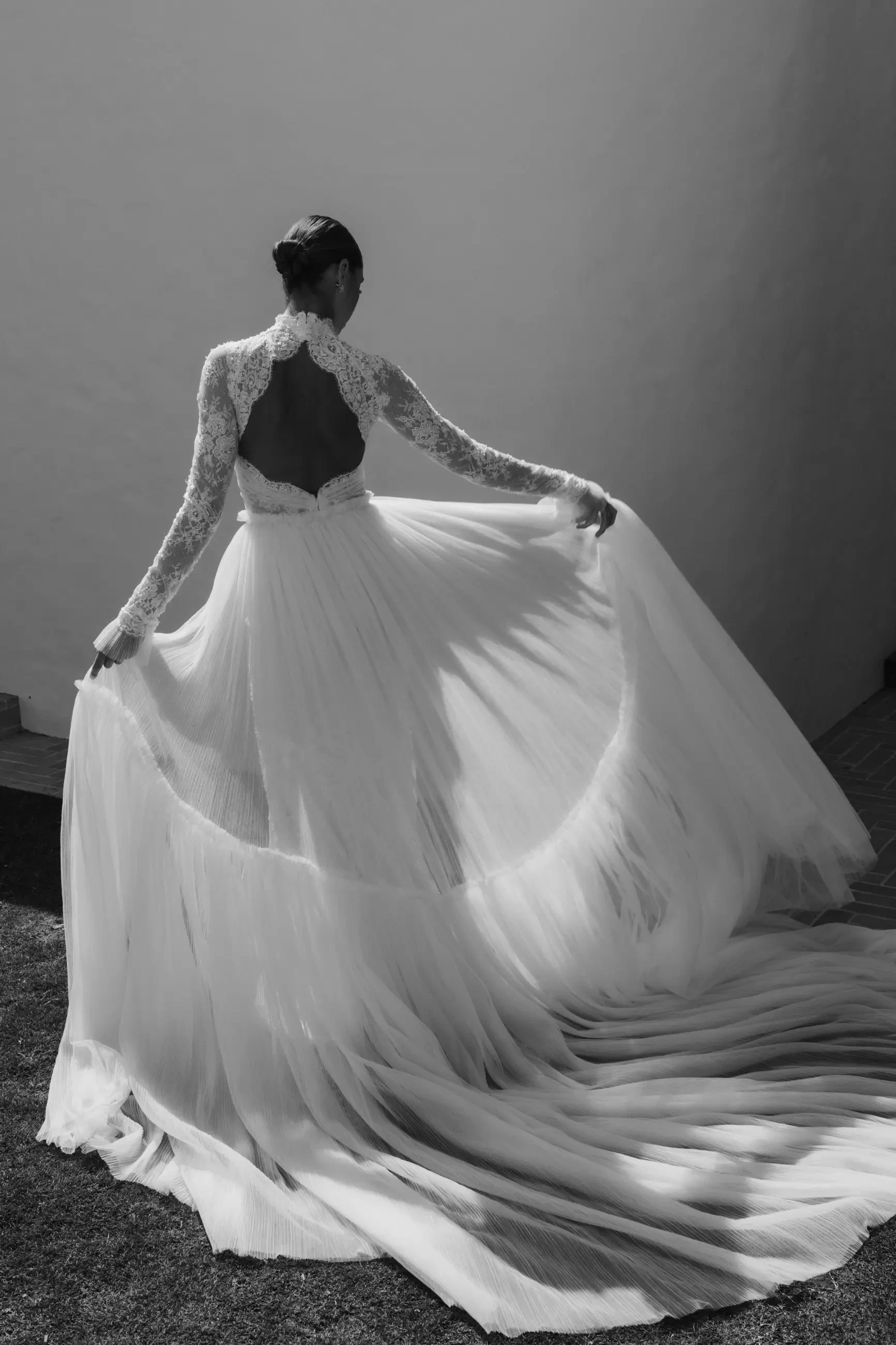 A figure in a flowing white gown with lace sleeves, turned away from the camera, showcasing the back of the dress and the train as it drapes elegantly to the ground. The image is in black and white.