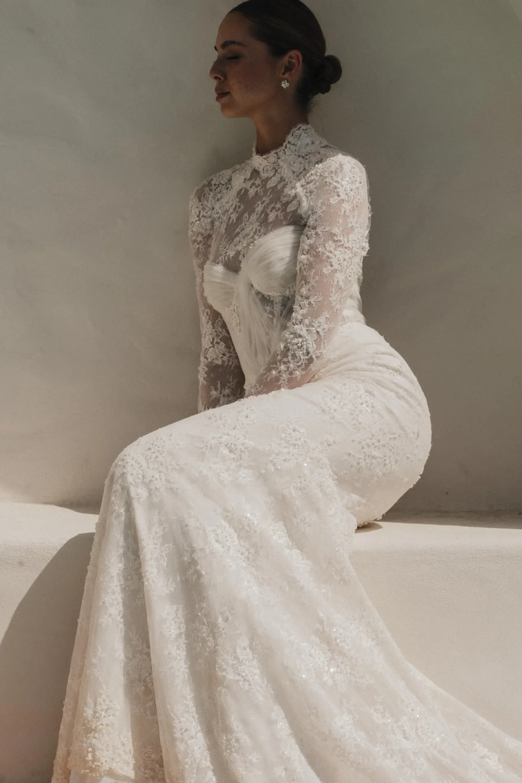 A woman wearing an elegant white lace wedding dress, sitting against a plain background.