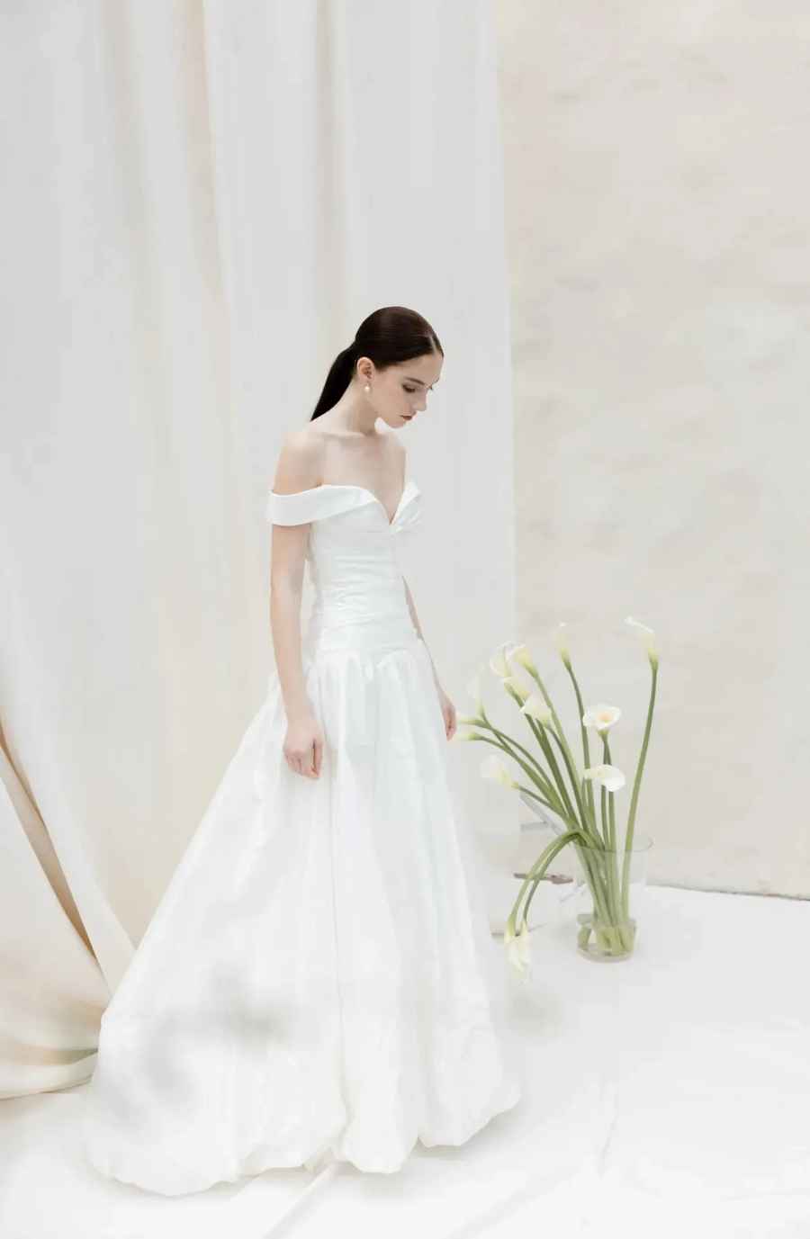 A person in a white off-the-shoulder dress standing next to a vase with flowers, surrounded by a light-colored background.
