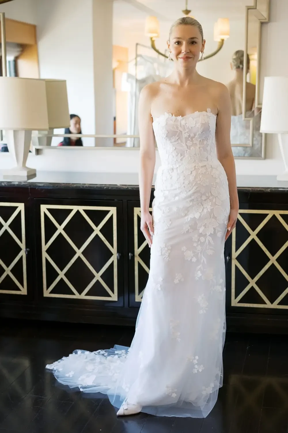 A woman in a strapless white wedding dress with floral details stands in front of a mirror, smiling.