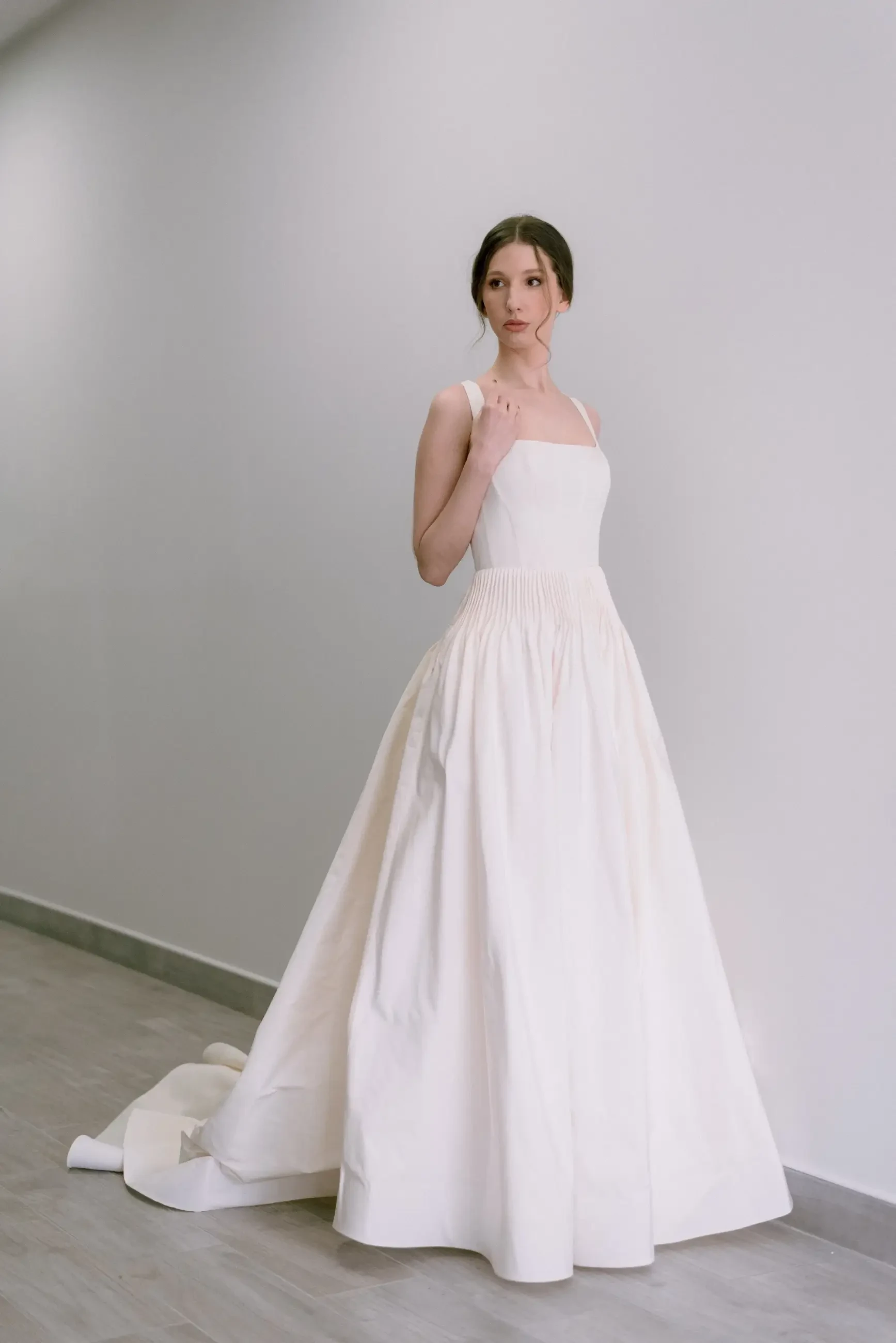 Woman in an elegant, white, sleeveless gown stands gracefully in a minimalistic setting. The dress features a full skirt, conveying a calm, poised mood.