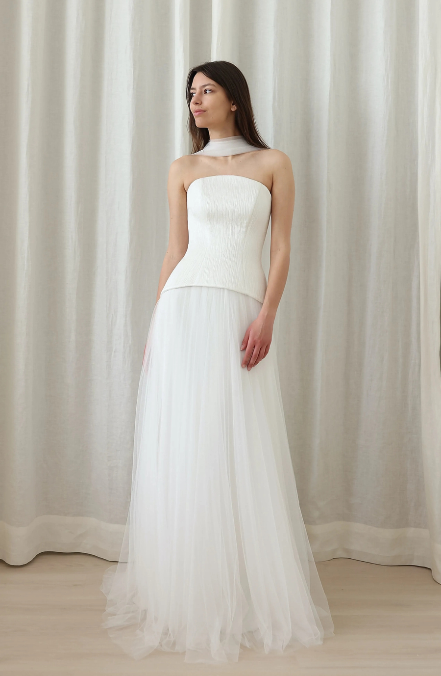 A woman wearing a strapless white gown with a flowing skirt, standing in front of a light-colored curtain.